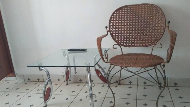 A small glass table with metal legs, a wicker chair with a woven backrest, and a tiled floor with a diamond pattern. - Dream House Hotel (Kisumu)