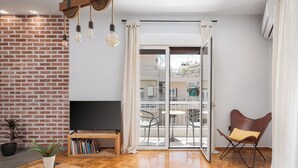 A modern living room with a brick wall, wooden flooring, a flat-screen TV, a bookshelf, a coffee table with a book, a potted plant, and a leather chair. - Acropolis Atmospheric Apartment (Athens)