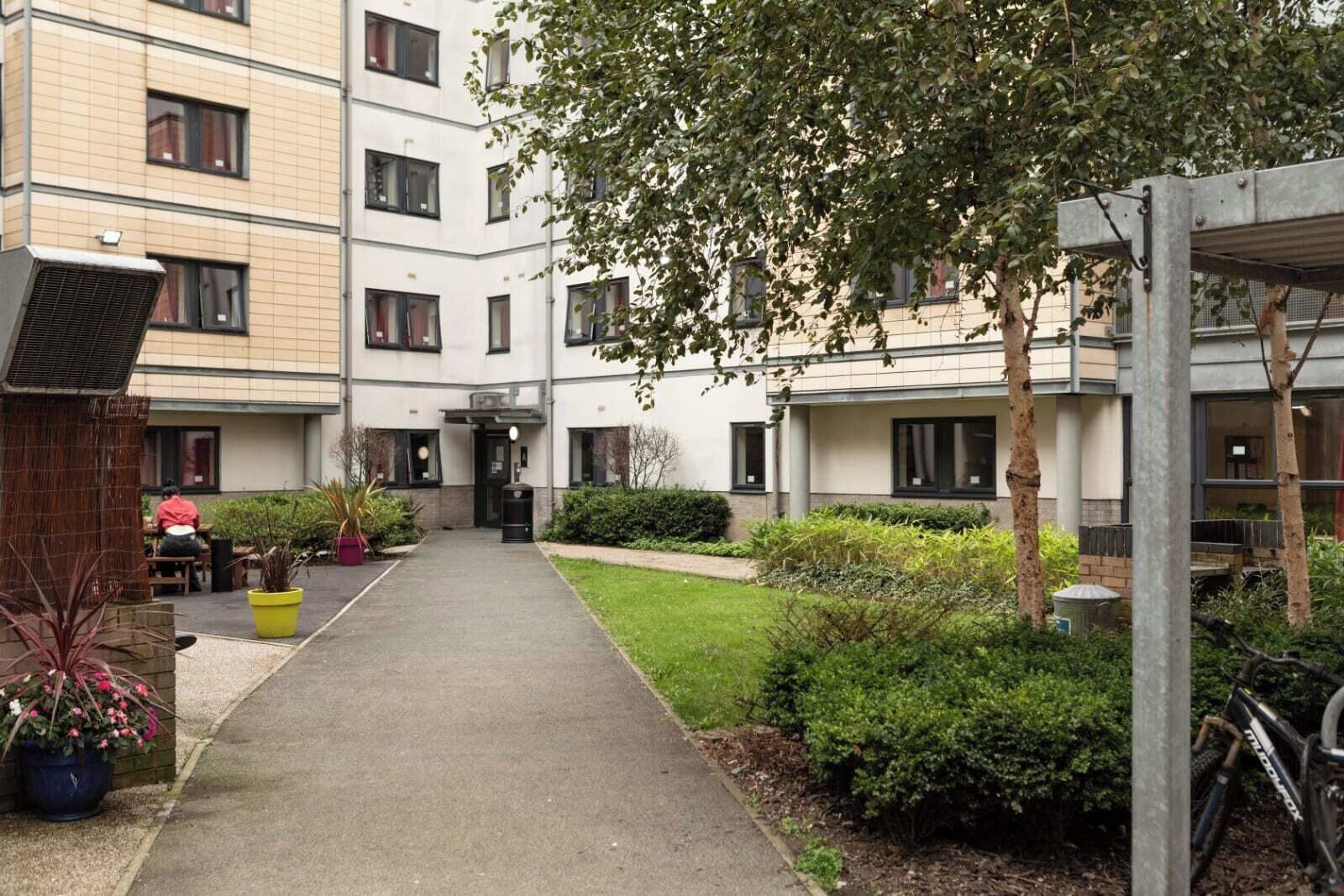 A courtyard with a pathway leading to a building entrance, surrounded by greenery and a parked bicycle.