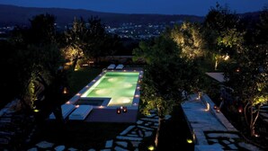 Vue nocturne d’une piscine rectangulaire, entourée d’arbres et bordée d’un chemin en pierre.
