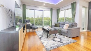 Un salón moderno con sofá, mesa de centro y sillas. Hay un escritorio con computadora y una planta. Amplias ventanas ofrecen vistas a la ciudad.