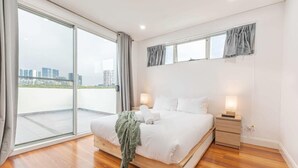 Un dormitorio moderno con una cama grande, una mesita de noche y vistas a un paisaje urbano a través de ventanas del suelo al techo.