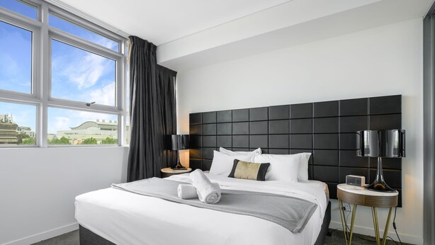 A modern hotel room with a large bed, a black headboard, a nightstand with a lamp, and a view of buildings outside. - Prime Location 1BD Apartment in Chatswood (Chatswood)