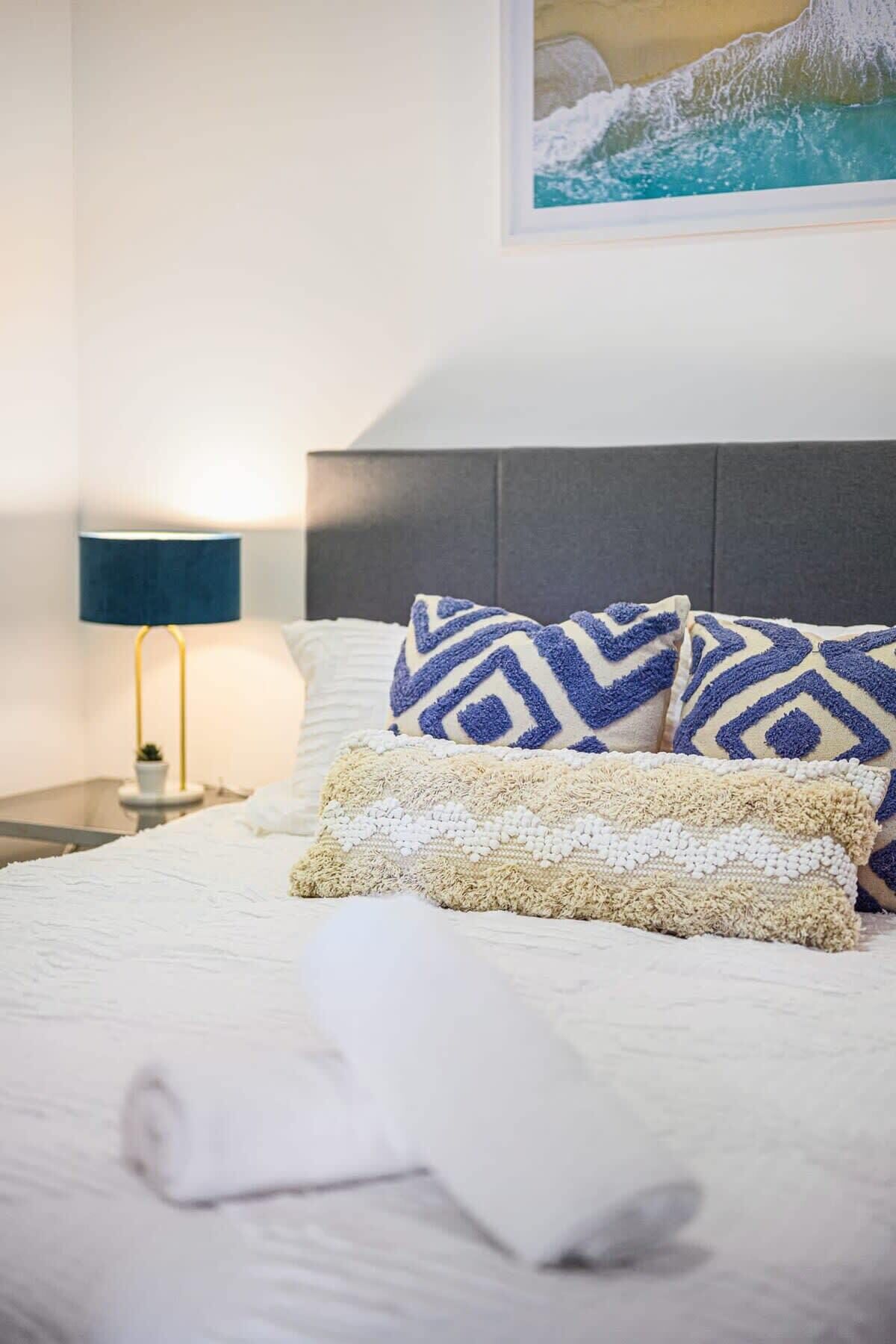 Ein ordentlich gemachtes Bett mit gemusterten Kissen, einer Lampe und einem Bild an der Wand.