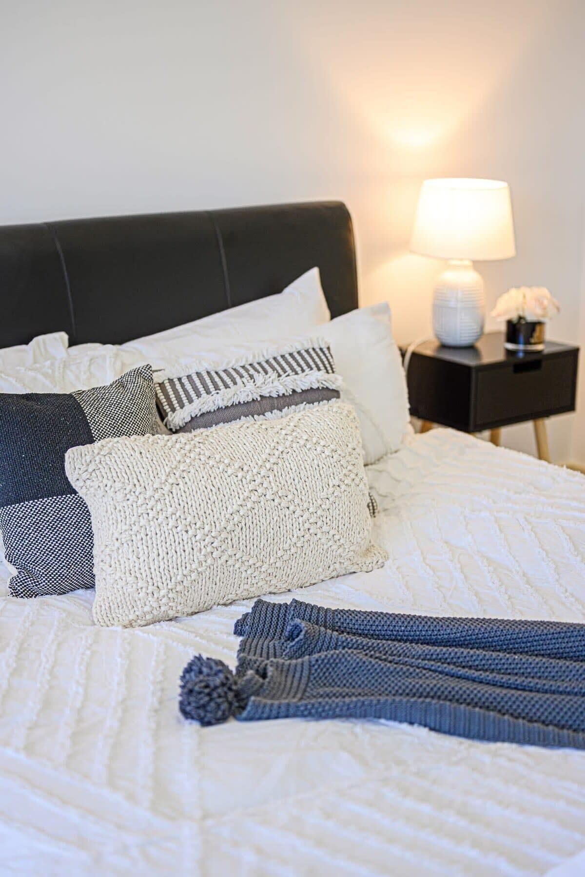 Ein ordentlich bezogenes Bett mit weißer Steudecke, mehreren Kissen und einer graublauen Decke. Ein schwarzer Nachttisch mit weißer Lampe und einer kleinen Vase mit Blumen steht neben dem Bett.
