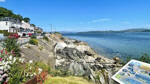 A coastal road with parked cars, a stone building, and a rocky shoreline. - Luxury Glamping Escape - Hot tub - West Coast Scotland (Innellan)