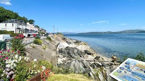 A coastal road with a stone wall, parked cars, and a flower bed.