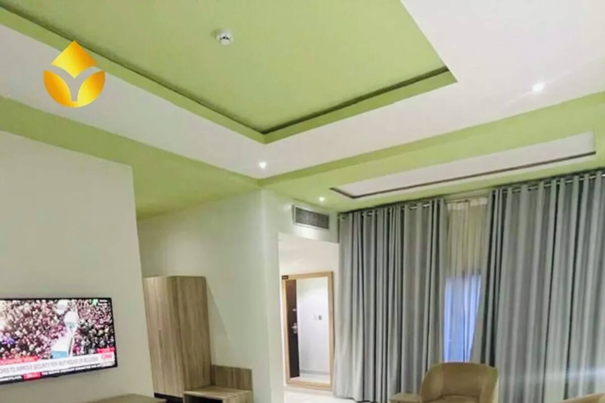 A modern hotel room with a large bed, a TV, a seating area with sofas and a coffee table, and a view of the outside through curtains.