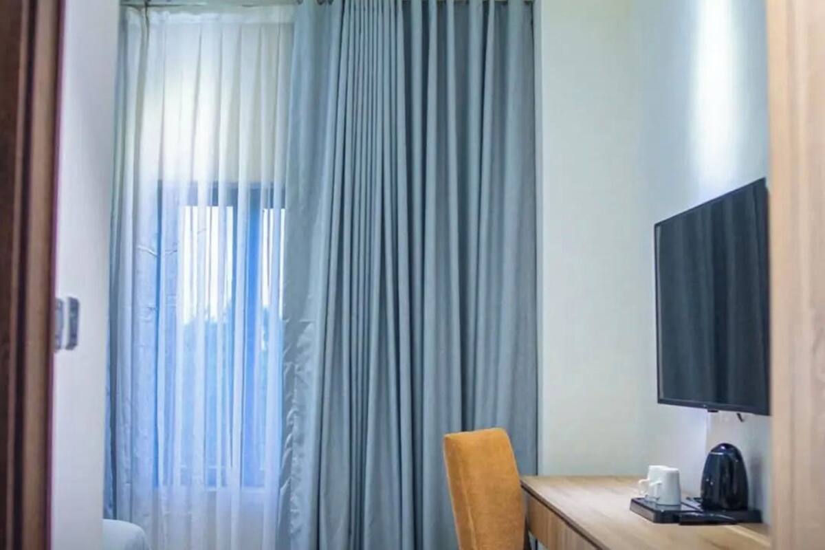 A hotel room with a large window covered by sheer curtains, a wooden desk with a television, and a chair.