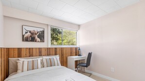 Une chambre à coucher avec une tête de lit en bois, un lit recouvert de draps blancs, un bureau, une chaise, une fenêtre avec des stores et un tableau encadré accroché au mur.