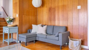 Un salon moderne avec un canapé d’angle gris, une petite table d’appoint, un luminaire suspendu et un panier en osier.