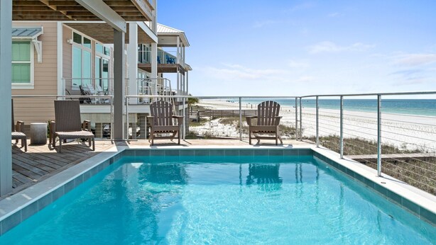 A swimming pool with a view of the beach, wooden deck chairs, and a building with balconies. - Bird of Paradise: Beachfront Views, Multiple Living Areas, Pet-Friendly (Mexico Beach)