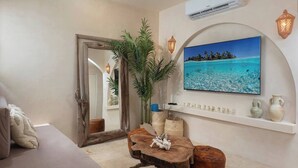 A living room with a large mirror, a wooden coffee table, a potted plant, and a wall-mounted TV displaying a tropical beach scene.