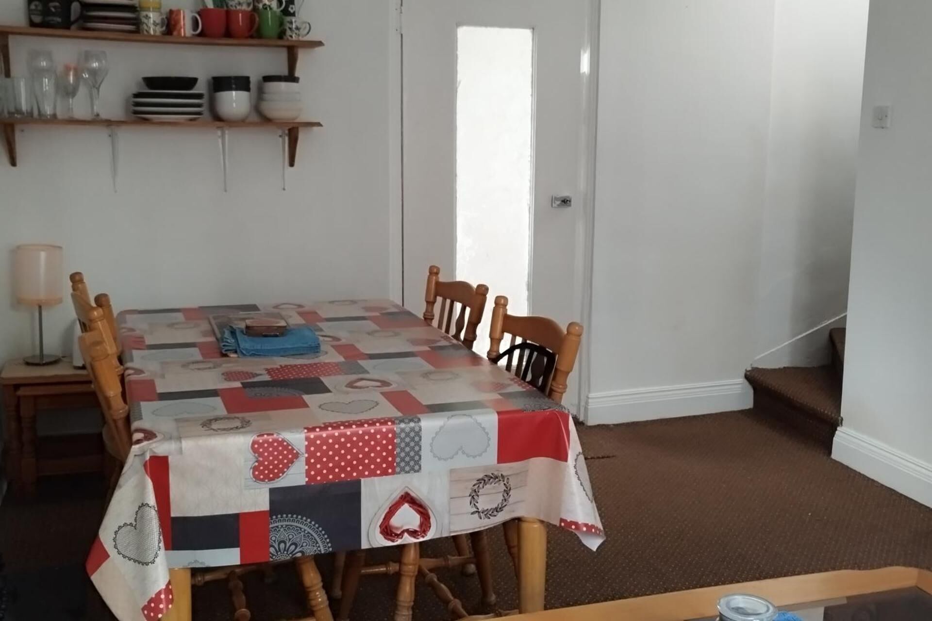 Uma área de refeições com uma mesa coberta por uma toalha de retalhos, cadeiras de madeira, uma luminária e prateleiras com pratos e copos.