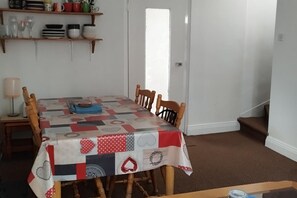 Un comedor con una mesa cubierta por un mantel de retazos, sillas de madera, una lámpara y estantes con vajilla y copas.