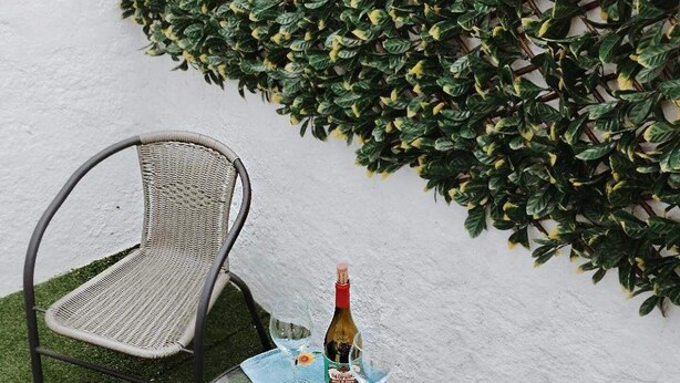 Een klein terras met twee rieten stoelen en een glazen tafel, een fles wijn en een glas op de tafel.