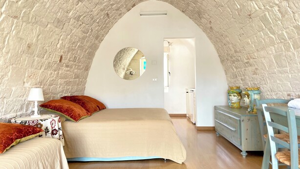 Une chambre à coucher avec un plafond voûté en pierre, un lit avec une literie rouge et beige, une table de chevet avec une lampe et une commode agrémentée d’objets décoratifs.