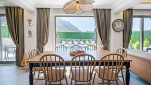 Espaço de refeições com mesa de madeira, cadeiras e vista para uma piscina e montanhas.