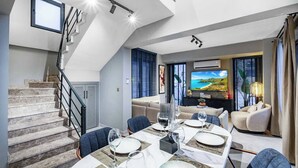 Una sala de estar moderna con comedor, una escalera y una gran pantalla de televisión mostrando un paisaje.