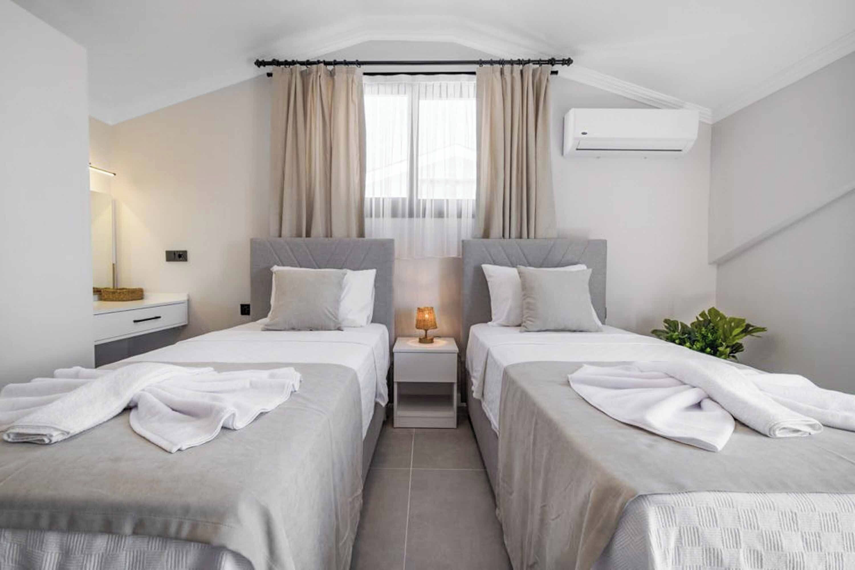 A hotel room with two beds, a bedside table with a lamp, a plant, and an air conditioning unit.