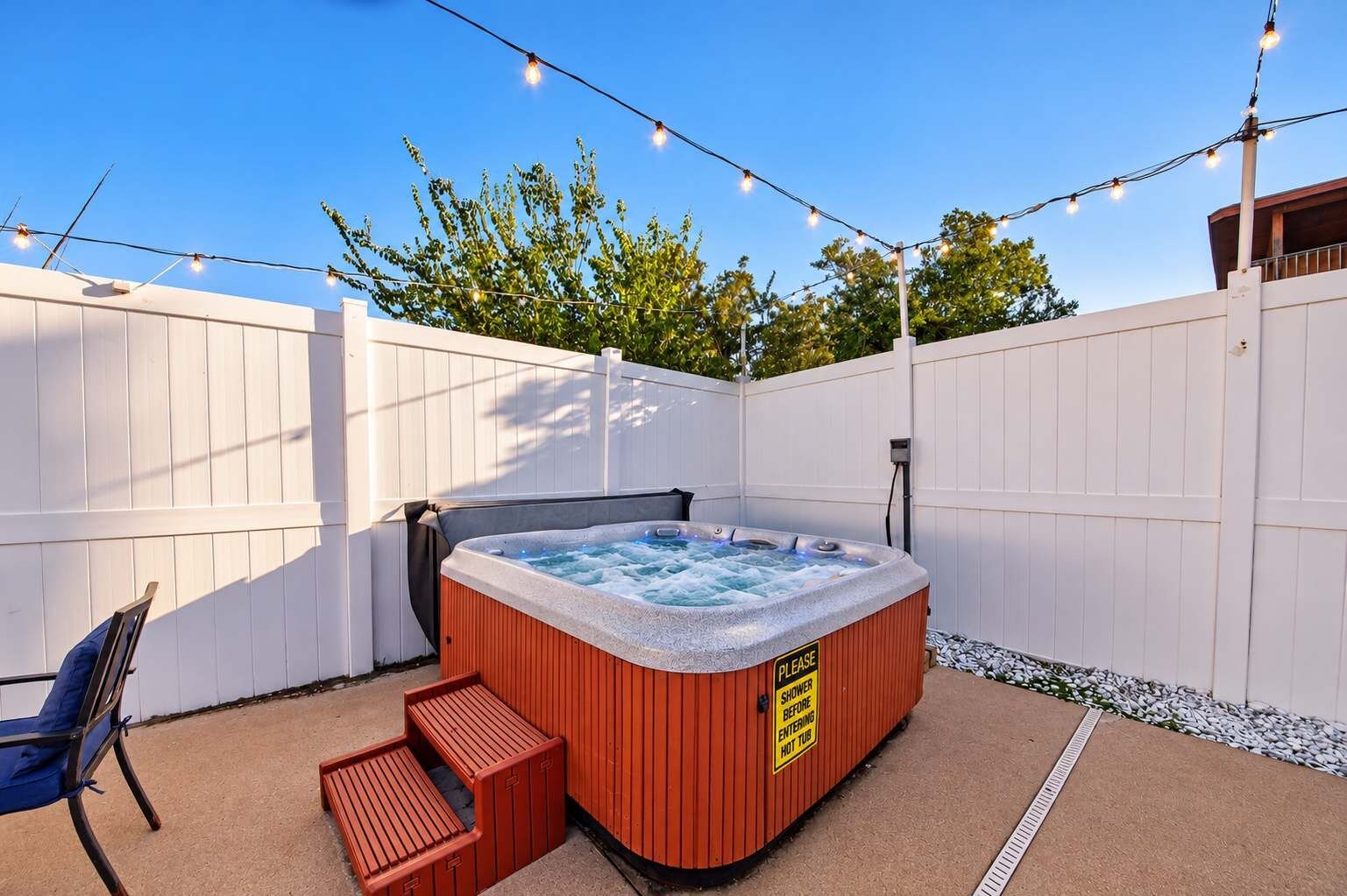 Un jacuzzi con un cartel que dice 'DUCHA ANTES DE ENTRAR AL JACUZZI', rodeado por una cerca blanca, con luces colgantes arriba.