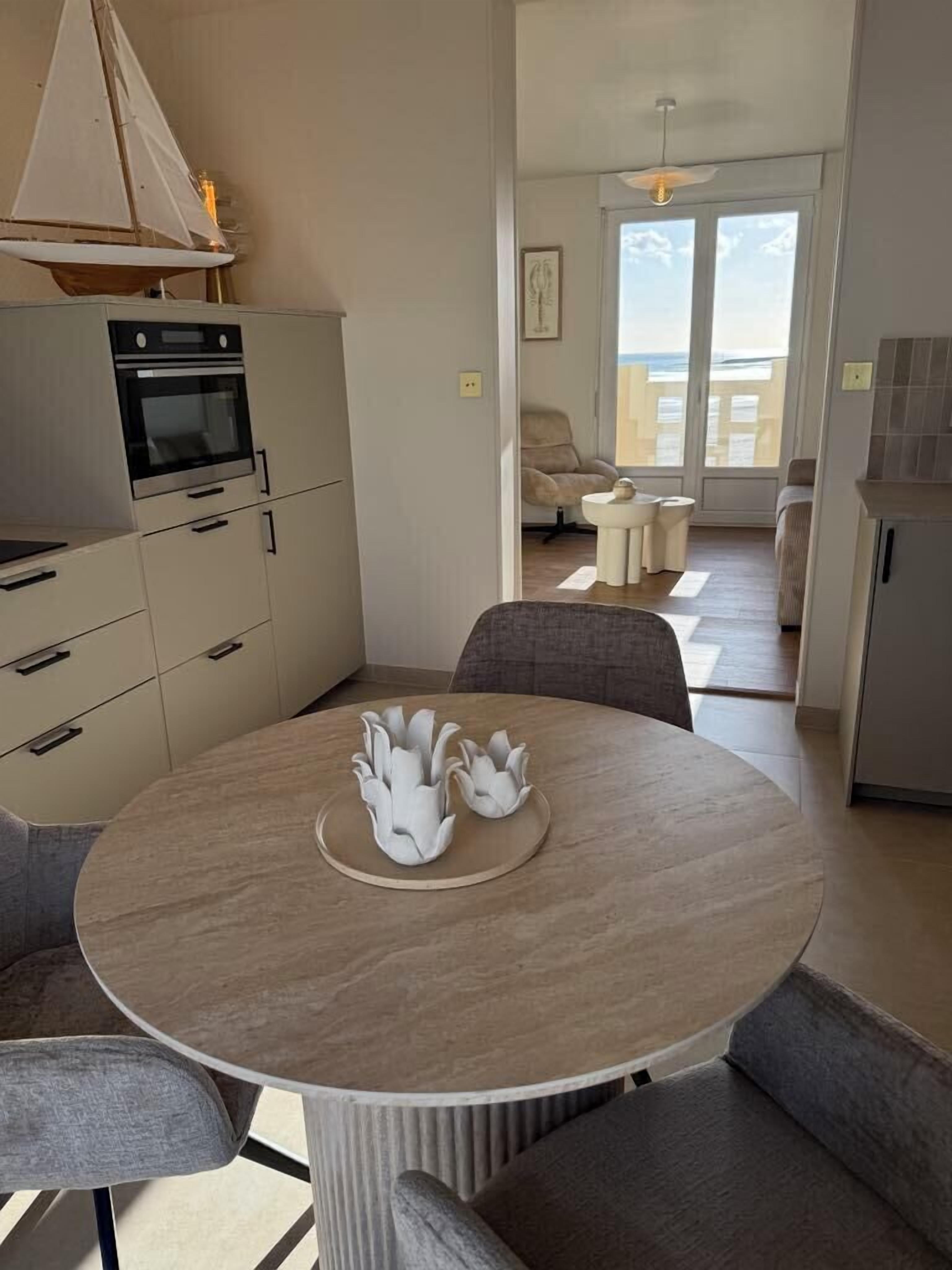 A modern kitchen with a built-in oven and cabinets, a round dining table with two chairs, and a view of the balcony through the open door.