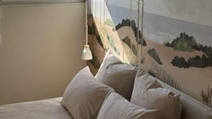 A bedroom with a bed, pillows, a wall mural, a window, and a hanging lamp.