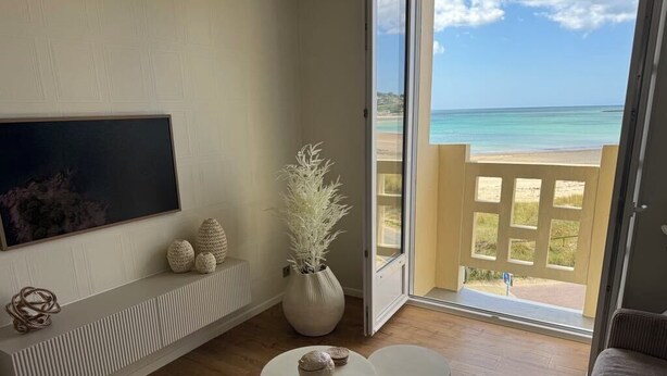 A living room with a large TV, two white side tables, a potted plant, and a view of the beach through a glass door.