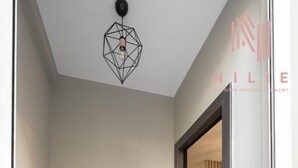 A hallway with a potted plant, a geometric light fixture, and a door leading to a room with a wooden floor and striped curtains.