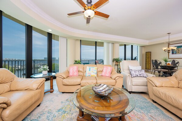 A spacious living room with a large window, a ceiling fan, a round glass coffee table, and a beige leather sofa.