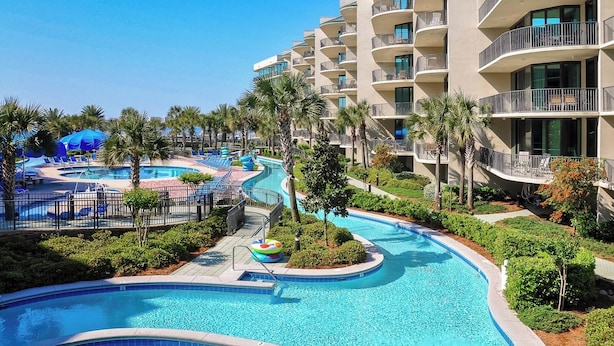 A resort with a large swimming pool, palm trees, and a multi-story building with balconies.