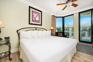 A hotel room with a bed, a ceiling fan, a framed picture, a nightstand, and a view of the outside.