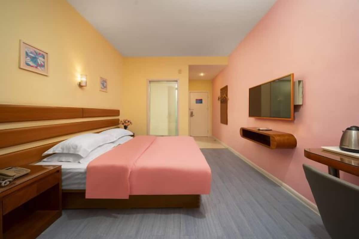 A hotel room with a bed, bedside table, a desk with a chair, a television mounted on the wall, and a door leading to another room.