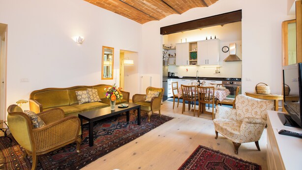 A living room with a sofa, chairs, a coffee table, and a TV. There is a kitchen in the background with cabinets and a clock. - Dimora Mattonaia: Cozy apartment with AC and WiFi (Firenze)