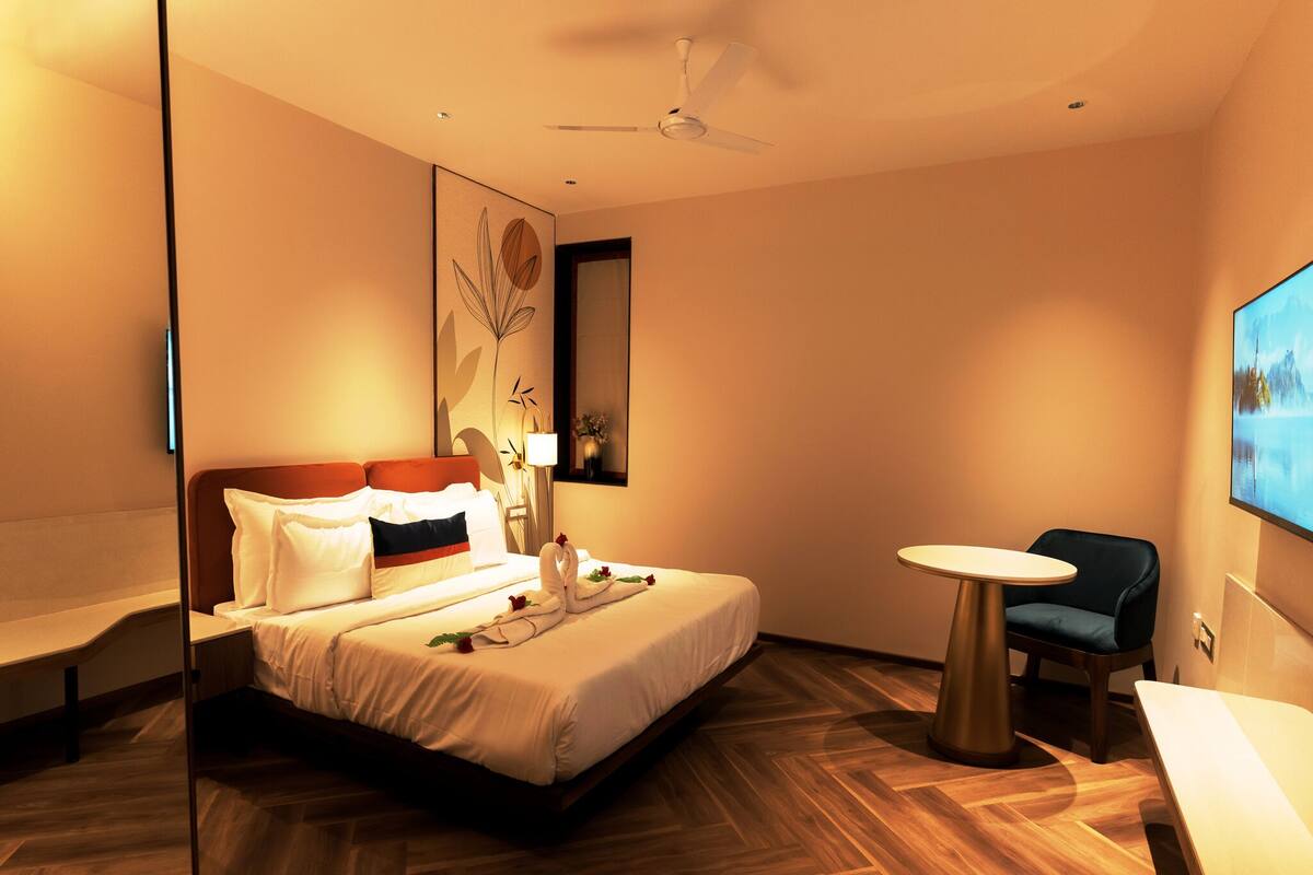 A hotel room with a bed, a round table, a chair, a ceiling fan, and a painting on the wall.