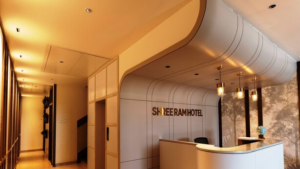 Ein moderner Hotel-Empfangsbereich mit einer gebogenen Rezeption, einem Schild mit der Aufschrift „SHREE RAM HOTEL“ sowie einem Empfängersitzbereich mit einer Weltkugel und einer Pflanze.