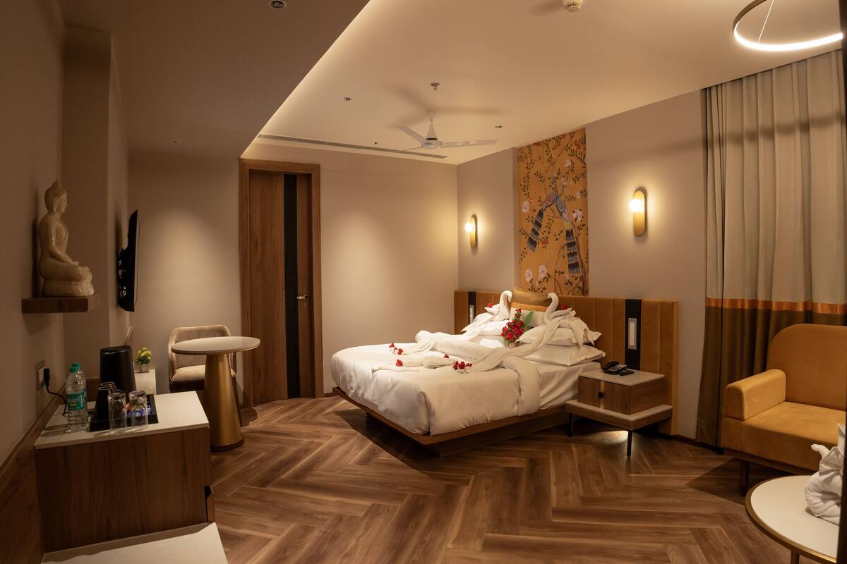 A modern hotel room with a wooden herringbone floor, a bed with white linens, a wooden nightstand, a desk with a chair, a sofa, and a wall-mounted TV.