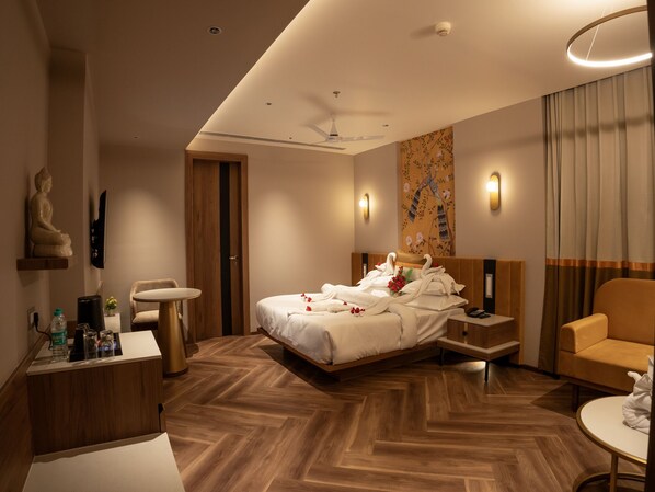 A modern hotel room with a wooden herringbone floor, a bed with white linens, a wooden nightstand, a desk with a chair, a sofa, and a wall-mounted TV. - Shree Ram Hotel (Beawar)
