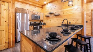 A wooden cabin interior with a kitchen featuring a stainless steel refrigerator, microwave, oven, and a large granite island with bar stools. - Gorgeous 3BD Near Creek! Hot Tub + FirePit + Games (Sevierville)