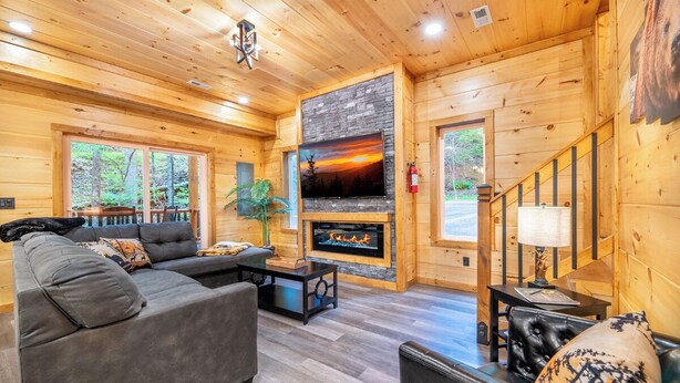 A cozy log cabin interior with a fireplace, a large TV, and a staircase. - Gorgeous 3BD Near Creek! Hot Tub + FirePit + Games (Sevierville)