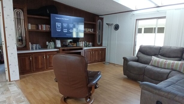 Uma sala de estar com poltrona de couro marrom, sofá cinza, armários de madeira e uma TV de tela plana mostrando um guia de conexão de rede.
