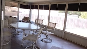 Uma área de varanda coberta com uma mesa redonda e seis cadeiras, janelas amplas e piso de concreto.