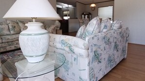Uma sala de estar com uma mesa de centro de vidro, luminária branca, sofá estampado e piso de madeira.