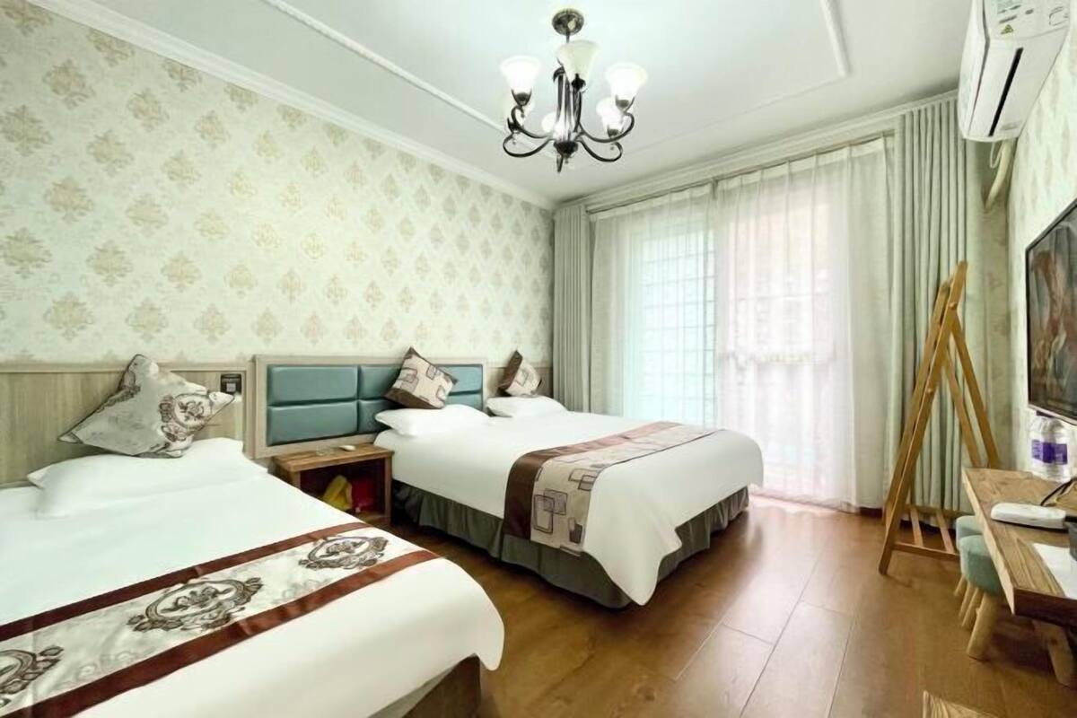 A hotel room with two beds, a wooden floor, a desk with a chair, a painting on the wall, and a chandelier.