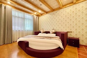 A bedroom with a curved bed, wooden ceiling beams, a patterned wallpaper, and a window with curtains.