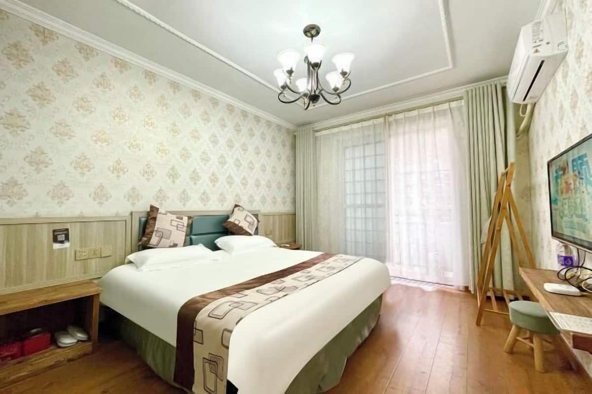 A hotel room with a bed, a desk with a computer, a chair, a wooden shelf, and a window with curtains.