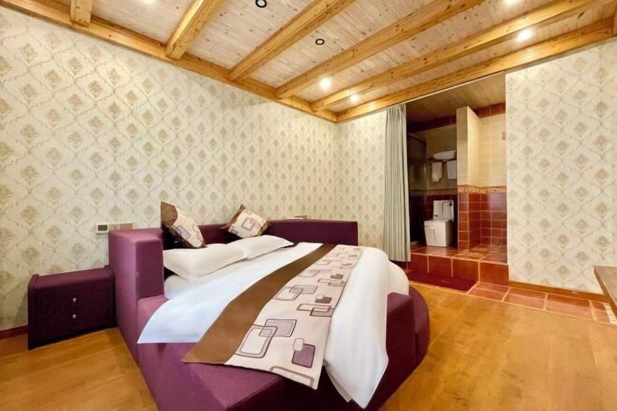 A bedroom with a wooden ceiling, patterned wallpaper, a bed with a brown headboard, and a bathroom visible through an open door.