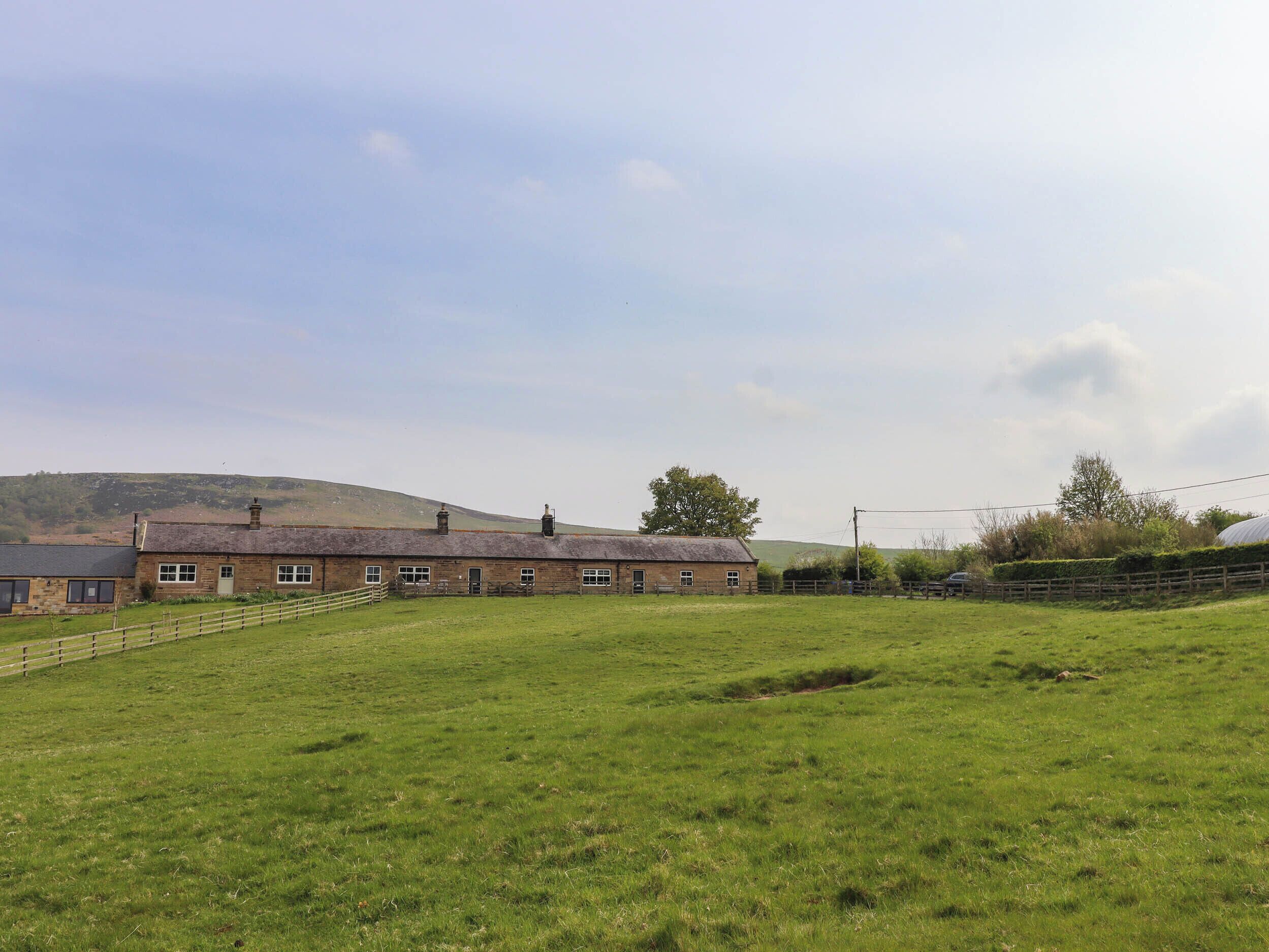 A rural property with a large open field, a stone building with multiple windows, and a hilly landscape in the background.