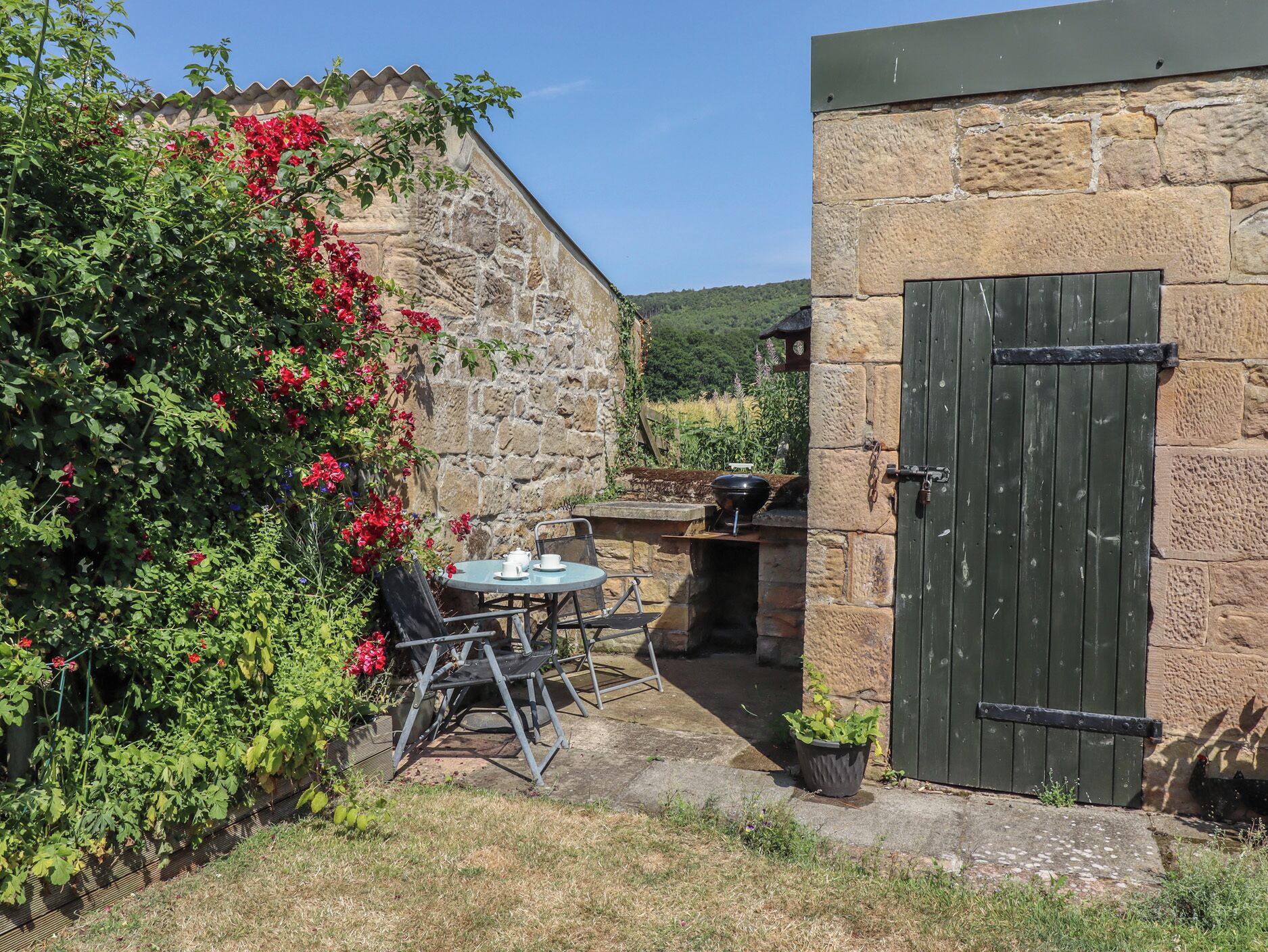 Un edificio de piedra con una puerta verde, una mesa con sillas y un jardín con flores rojas.