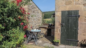 Un edificio de piedra con una puerta verde, una mesa con sillas y un jardín con flores rojas.